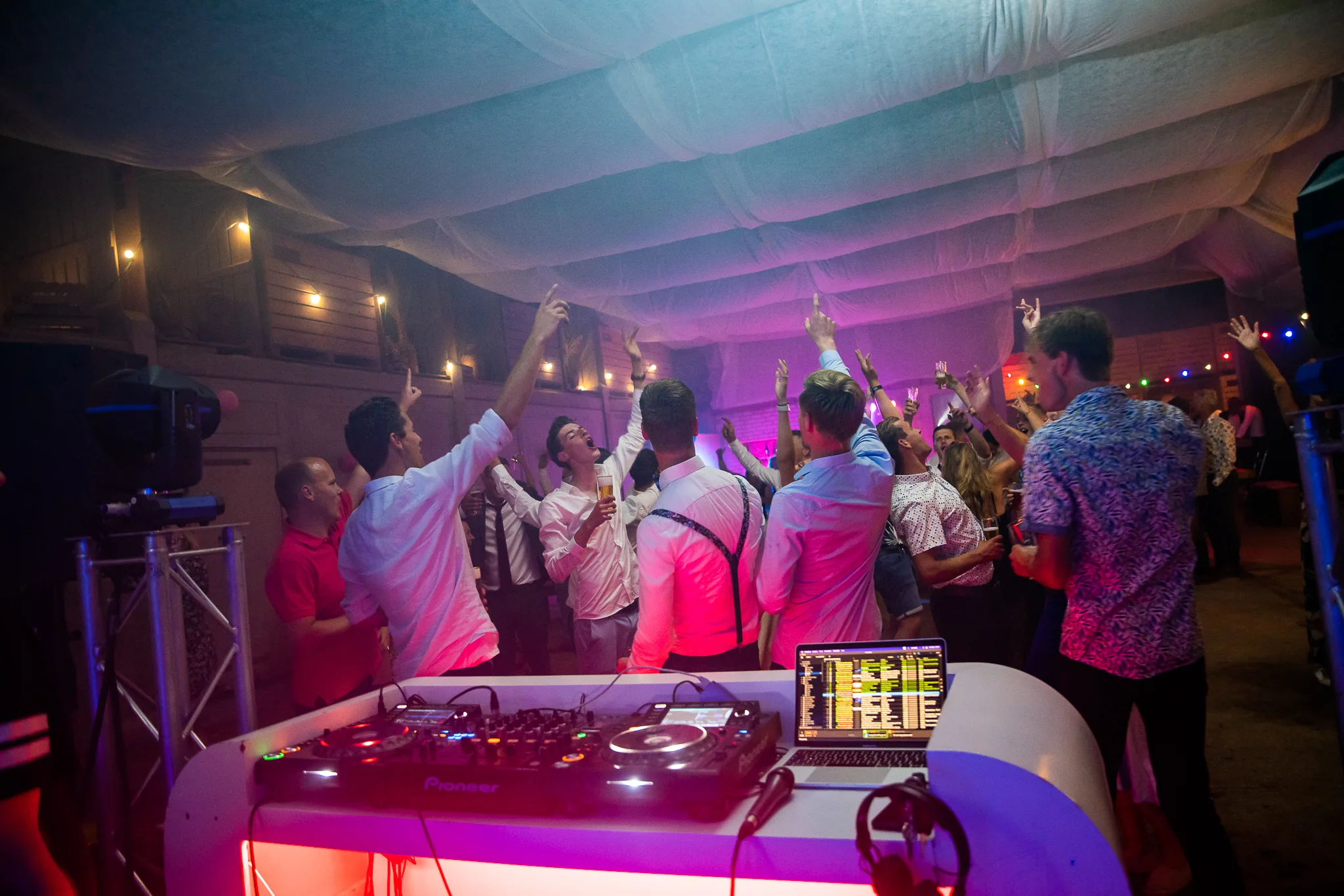 Bruiloftsgasten met de handen in de lucht voor een witte DJ-booth in een feesttent