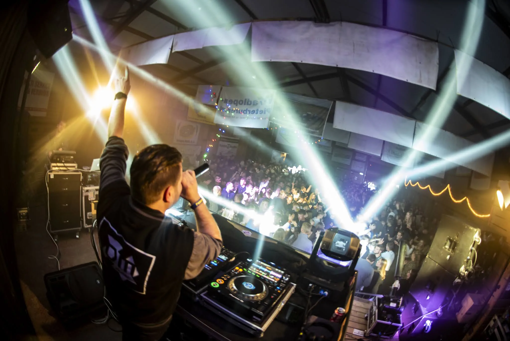DJ met de arm omhoog achter de booth en witte lichtbundels boven een volle zaal
