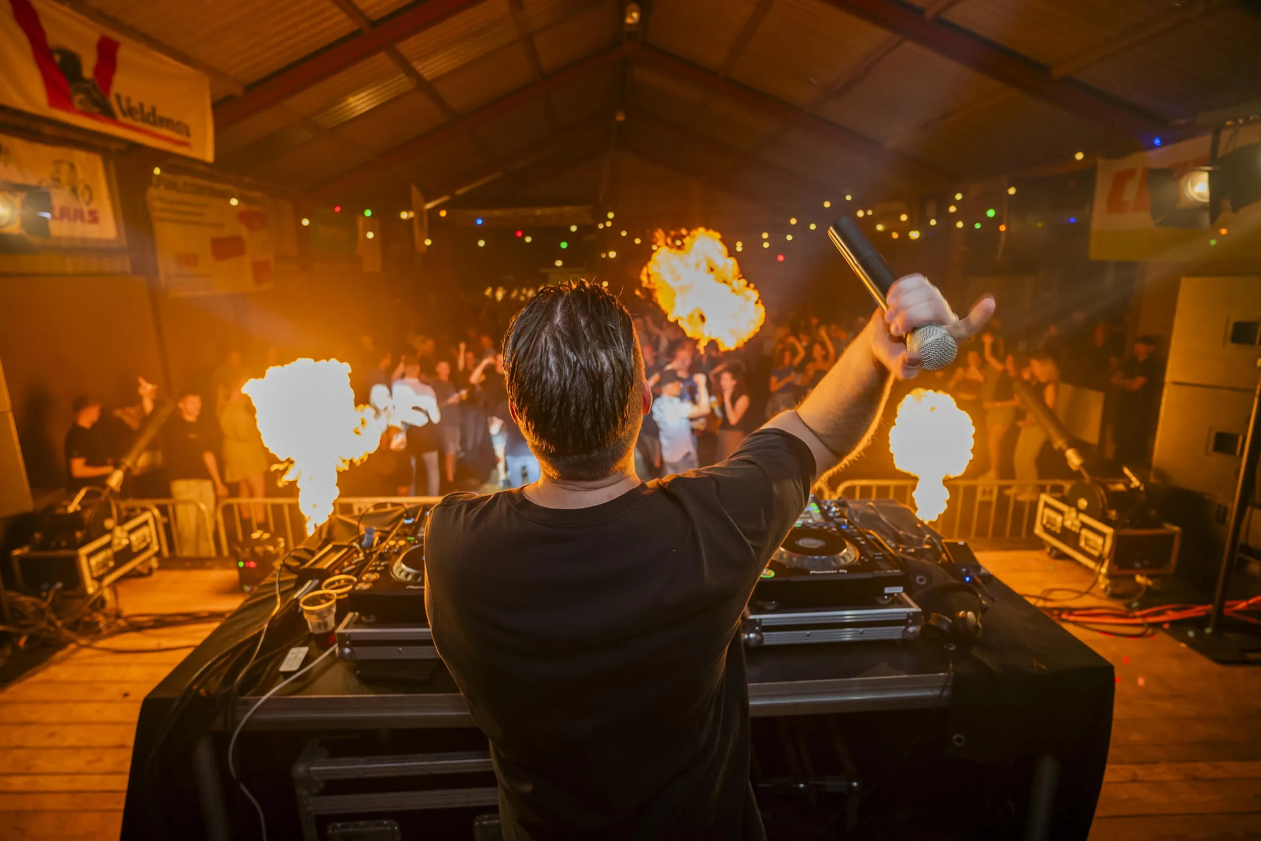 DJ met de microfoon omhoog achter de booth, met vlammen en publiek ervoor